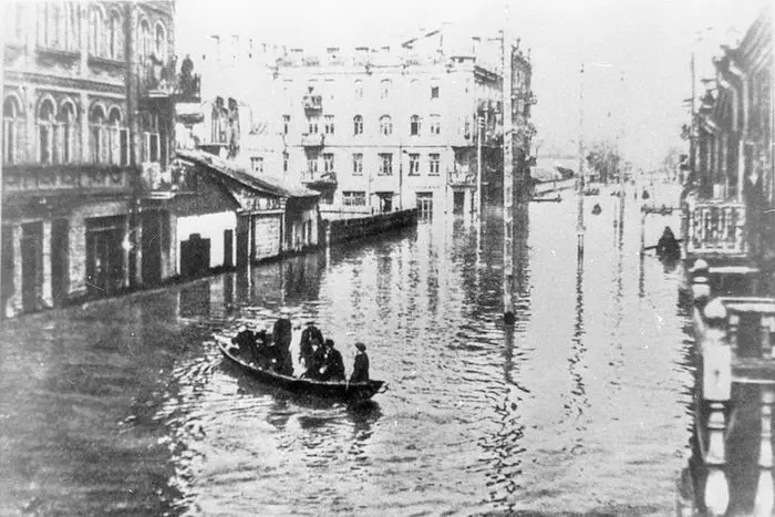 A historical black-and-white photograph showing a severe flood in the Podil district of Kyiv. People are being transported in a small wooden boat down a completely submerged city street lined with multi-story pre-revolutionary buildings.