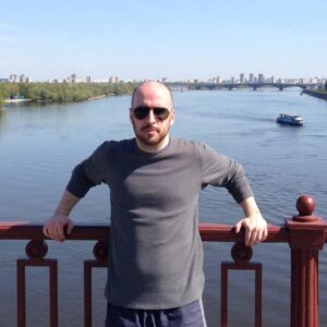 A portrait of Sean, the founder of Vestor Estate, wearing sunglasses and a grey long-sleeved shirt, leaning against the red railing of the Pedestrian Bridge in Kyiv. The background features a wide, sunny view of the Dnipro River, Trukhaniv Island, and the Kyiv city skyline under a clear blue sky.